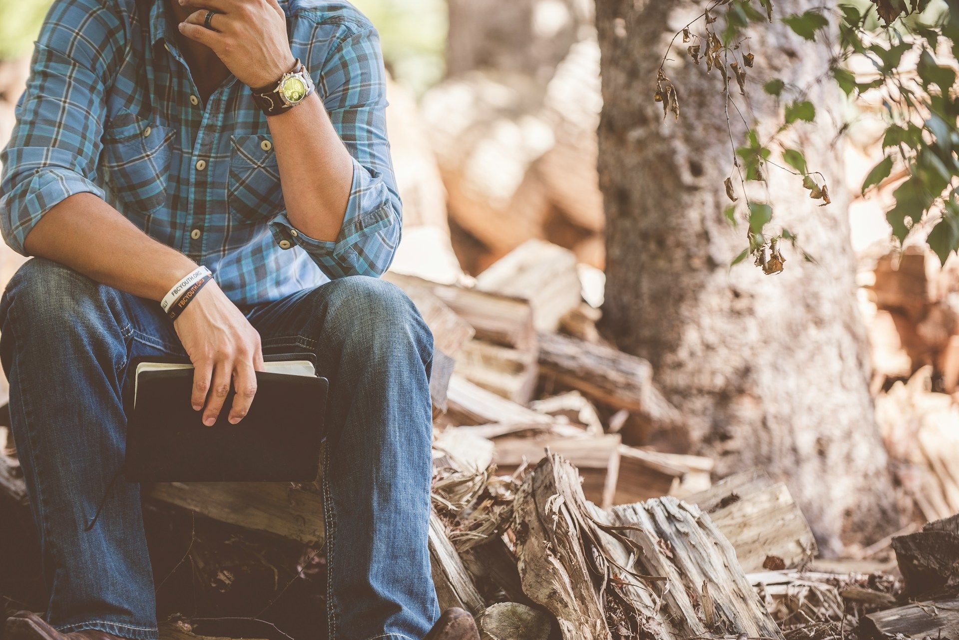 Photo of a man lost in thought, sitting on a pile of wood next to a tree.