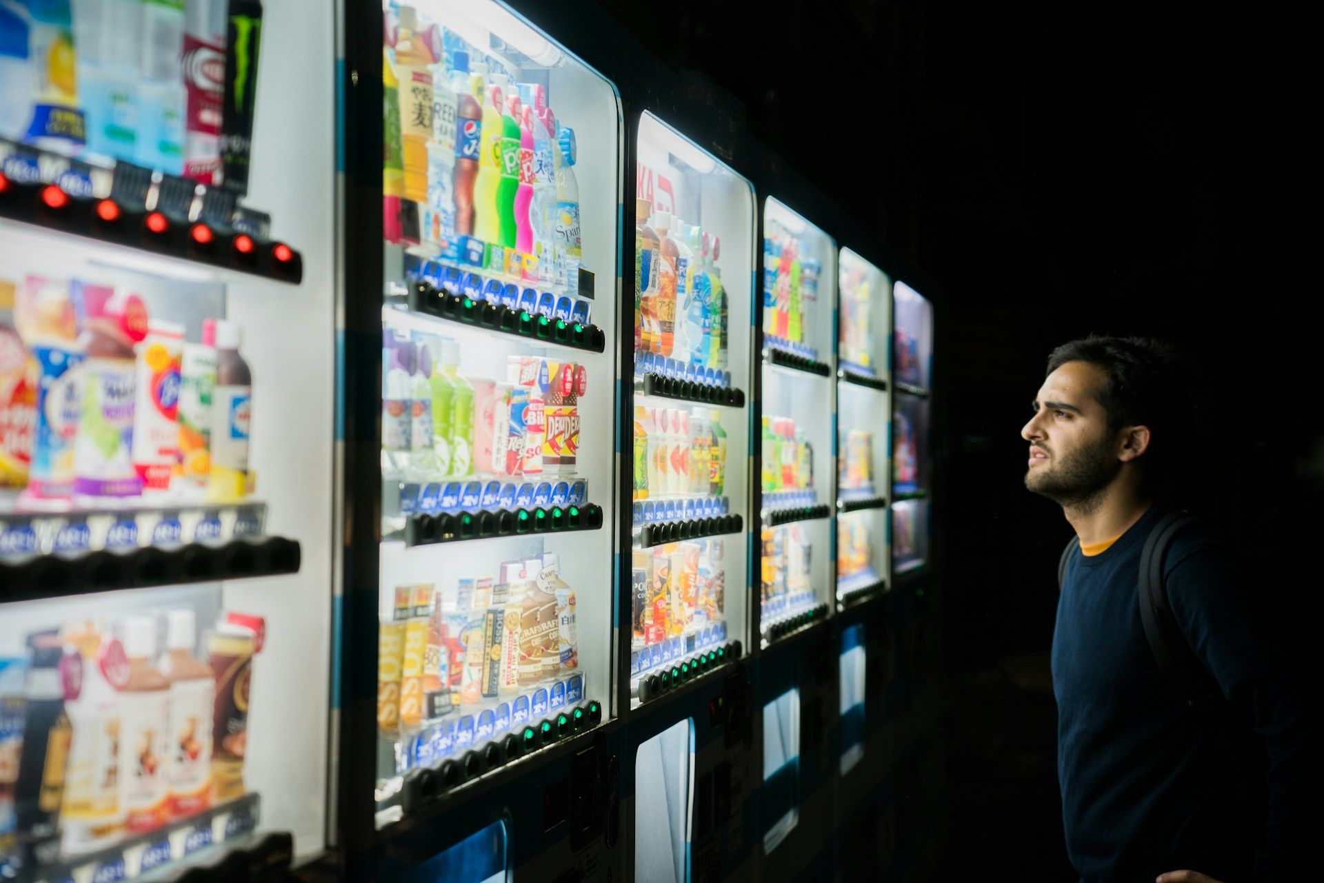 Man looking at a line of drink bottle vending machines in Japan, seemingly undecided which drink to buy.