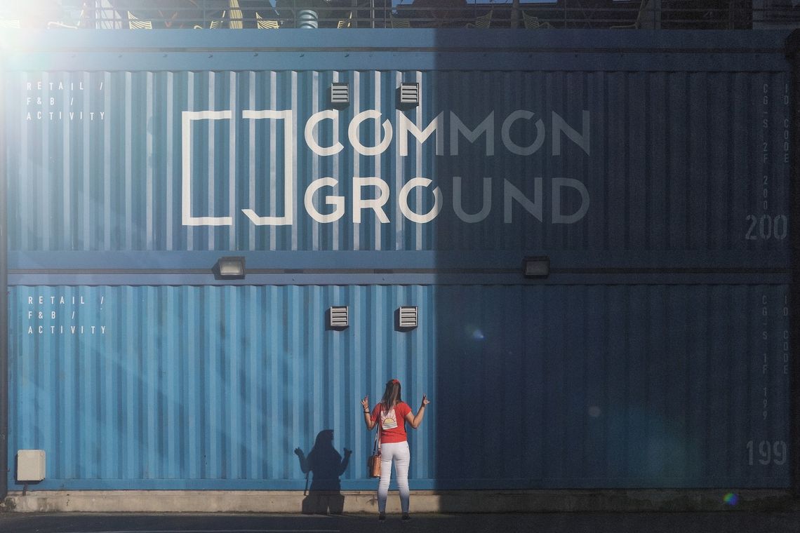 Person with long hair facing two stacked blue transport containers. On the top one is written: "Common Ground"